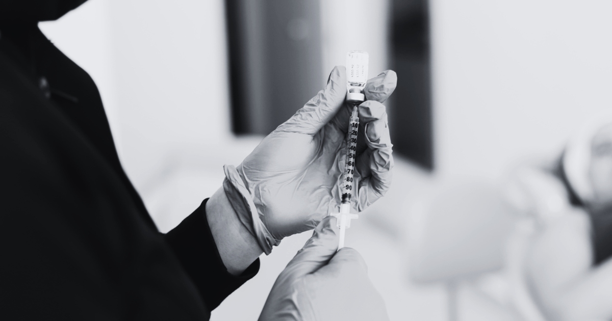 A person wearing disposable gloves, holding a syringe and a small vial.