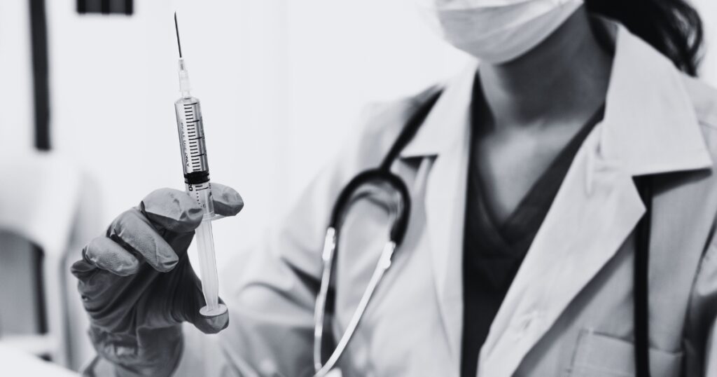 Black-and-white close-up of a medical professional in a lab coat and mask holding a syringe upright, with a stethoscope visible around their neck.