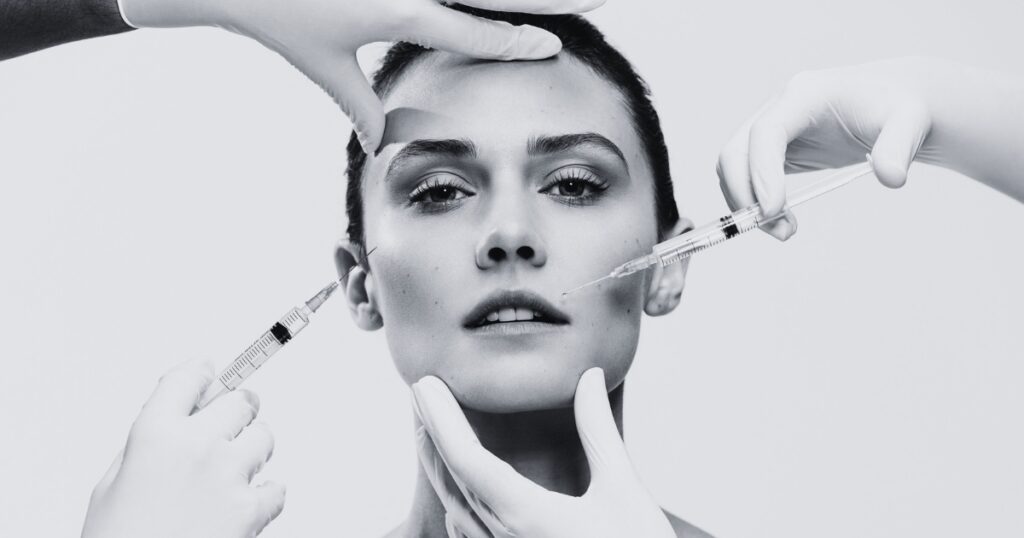 Black-and-white close-up of a woman facing forward while multiple gloved hands hold her face and administer cosmetic injections to her cheeks and temple against a plain background.