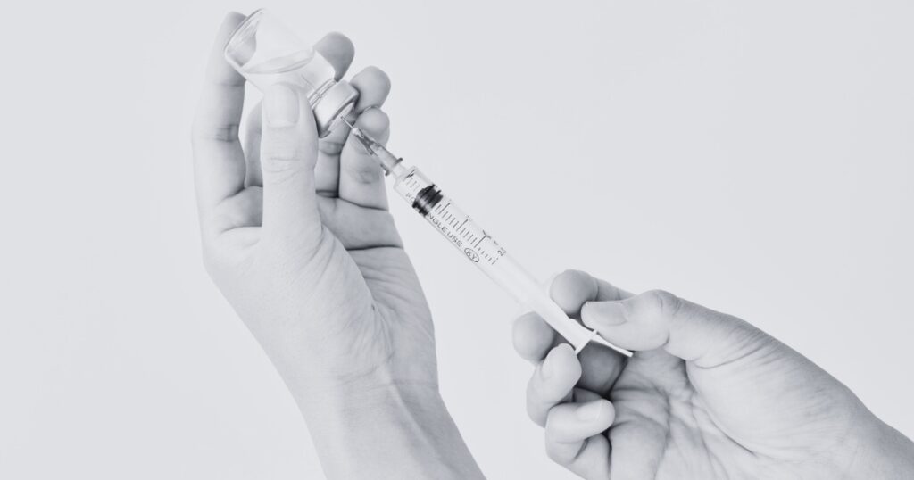 Black-and-white close-up of gloved hands drawing liquid from a small vial into a syringe, emphasizing a sterile medical or cosmetic preparation.