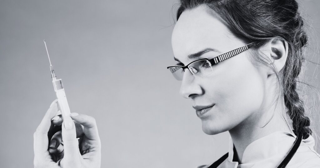 Black-and-white close-up of a medical professional wearing glasses and gloves, carefully holding and examining a syringe.