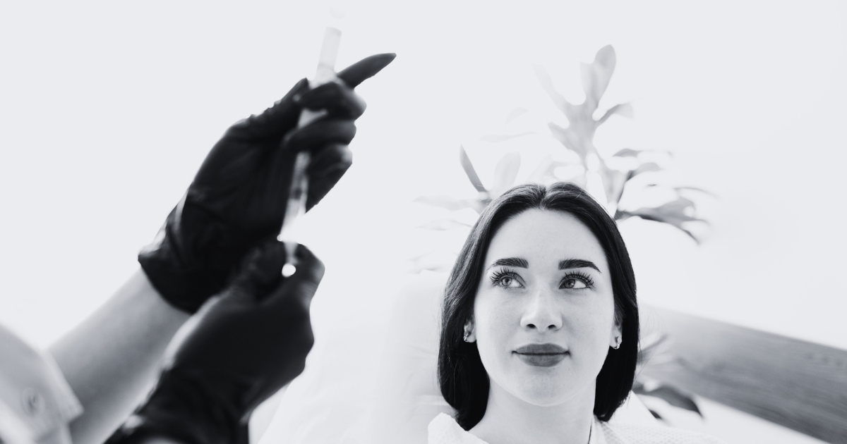 A woman looks upward while a medical professional wearing black gloves holds a syringe, preparing for a cosmetic injection procedure in a clinical setting.