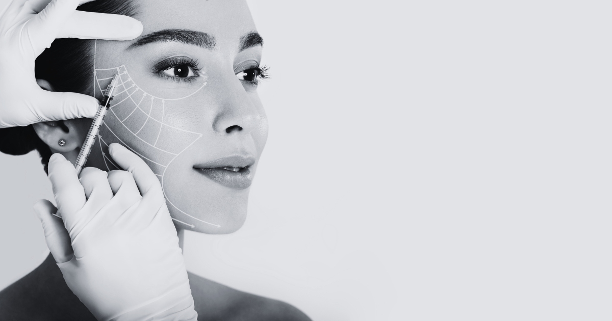Close-up of a woman’s face with facial mapping lines while a medical professional wearing gloves holds a syringe, preparing for a cosmetic facial injection procedure.